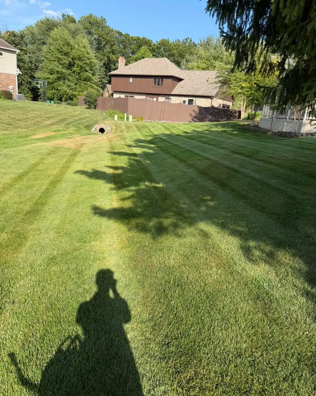 Mowing stripes running down a gentle slope toward a culvert pipe, with a brown timber house beyond