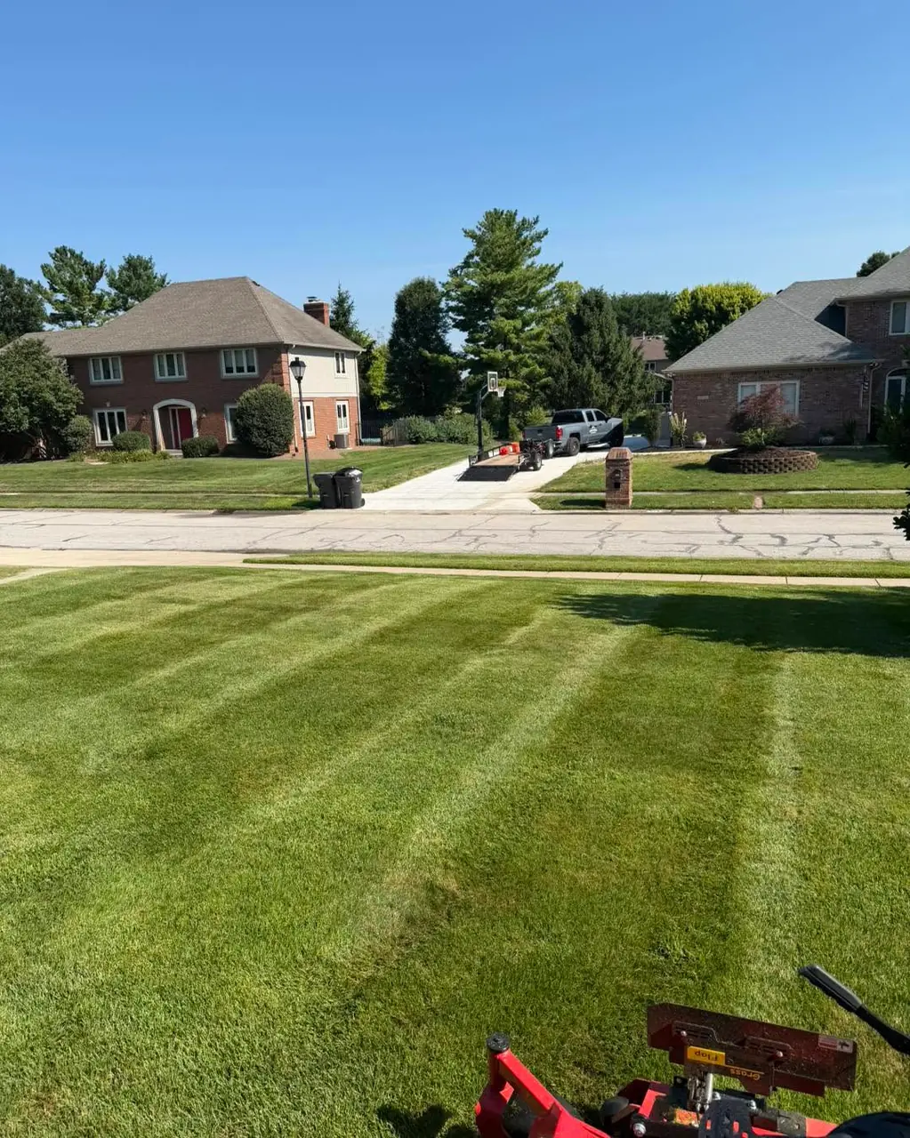 A striped front lawn looking out across the street to brick homes, with the mower deck in the bottom of the frame